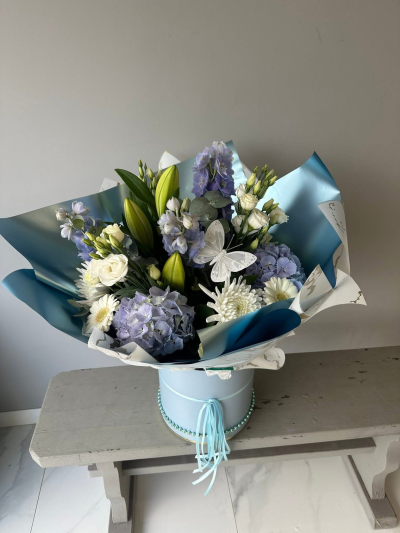 Azure Elegance - A beautifully handcrafted arrangement combining fresh seasonal blooms in soothing blue and white shades with abundant greenery for a soft, luxurious finish. Presented in a premium hat box and accented with a delicate butterfly, this gift is ideal for celebrating life's most precious moments.